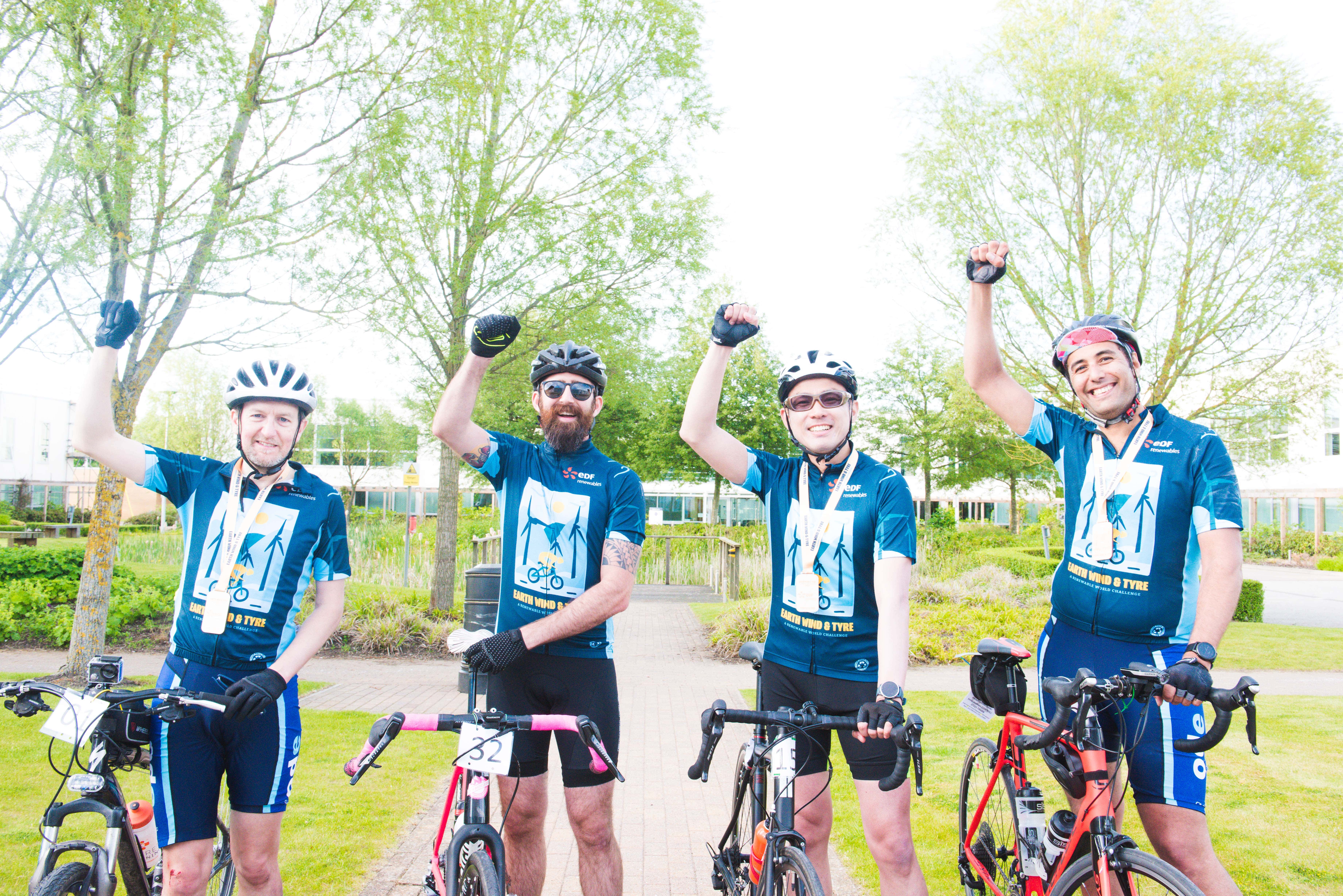 Four Earth Wind & Tyre cyclists standing next to their bikes with their fists held up in the air in celebration