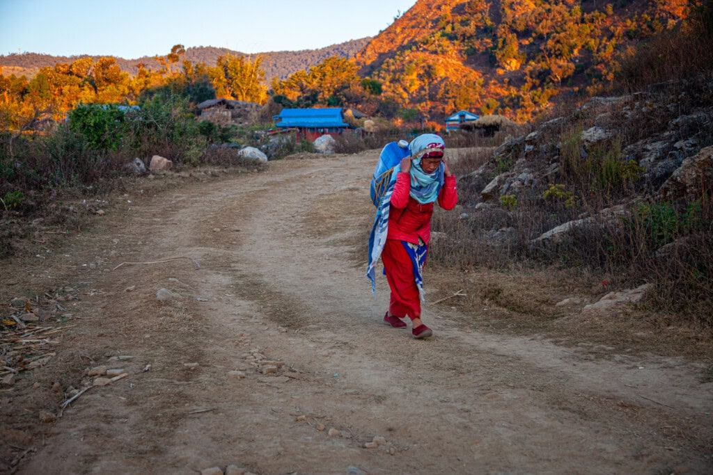 A woman is walking along a dirt path, carrying a heavy vessel of water on her head. In the background, you can she she is in a mountainous rural area.