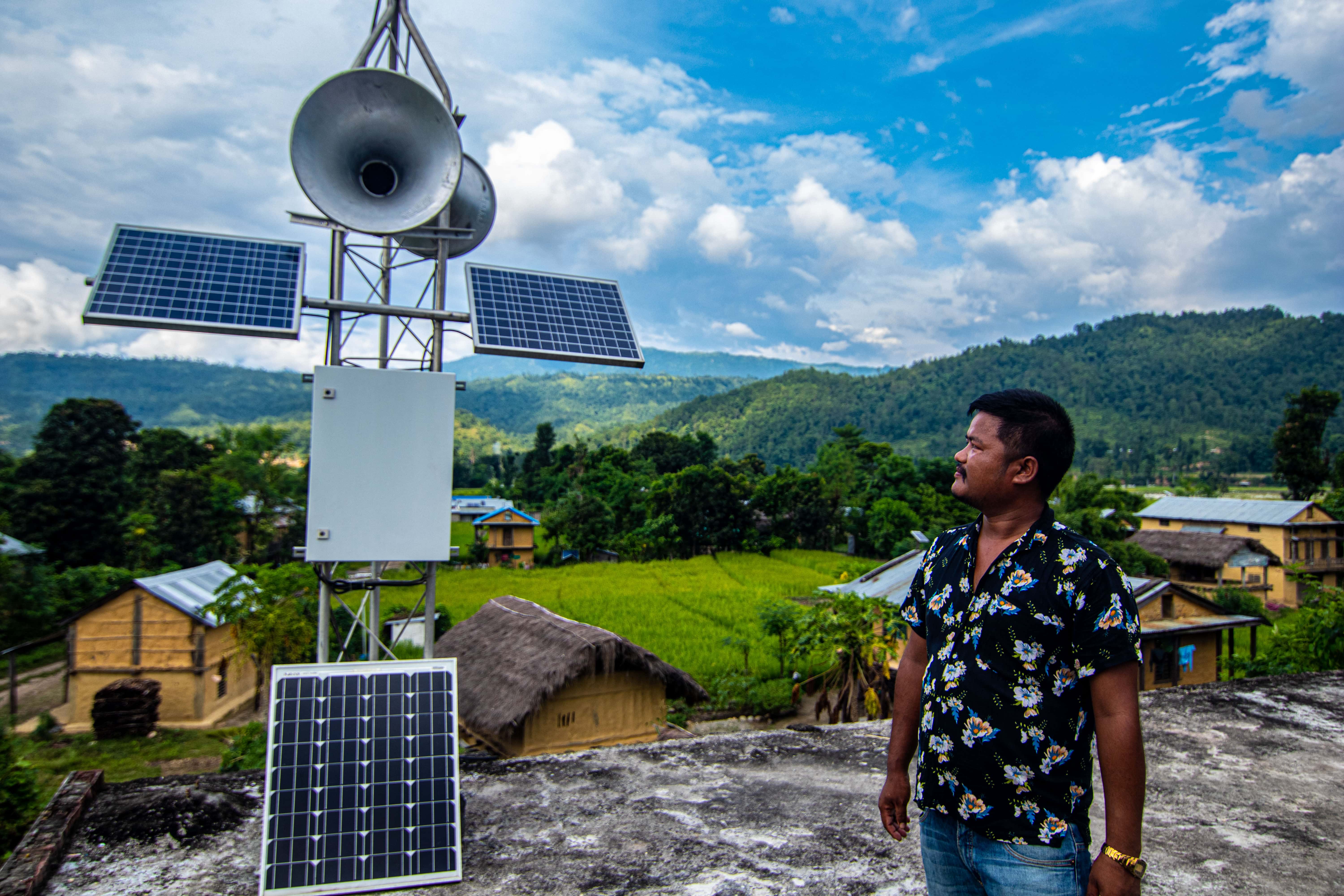 A Nepali man standing on a rooftop, looking at a mounted structure with solar panels and sirens - these are part of a solar-powered early warning system