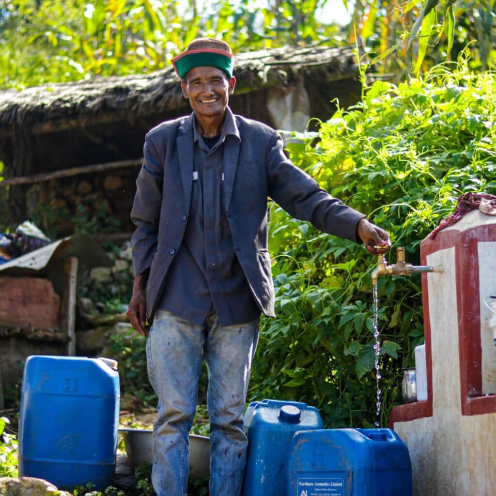 A Nepali man is standing in a rural landscape at a tap stand, with water running into a water vessel and many other water vessels around him. He is beaming at the camera.