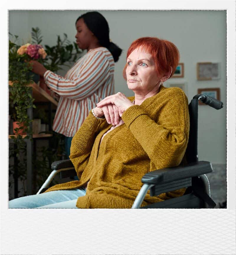 Elderly woman in wheelchair with caregiver arranging flowers in background.