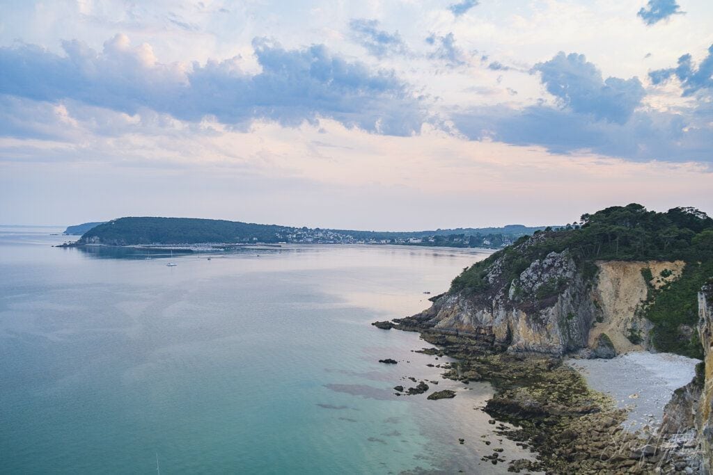 A lush coastal landscape featuring a rocky shoreline, green cliffs, and calm sea waters under a partly cloudy sky during sunset.