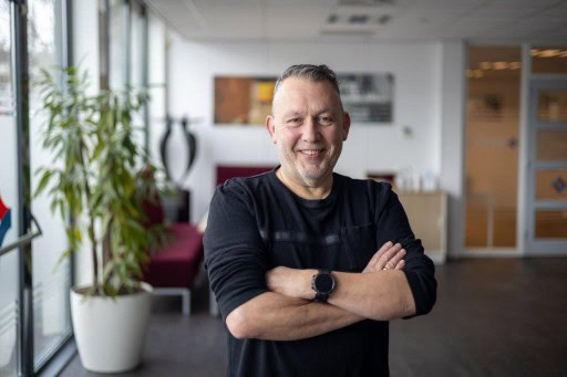 Rense Sjoers – Dutch hobbyist photographer ShiftyNL Photography, Professional man smiling confidently in a modern office environment, wearing a black shirt and smartwatch, with natural light and contemporary decor in the background.