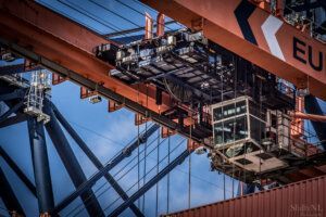 High-angle view of a large industrial crane lifting shipping containers, showcasing detailed machinery, steel structure, and blue sky background. Perfect for capturing industrial photography moments.