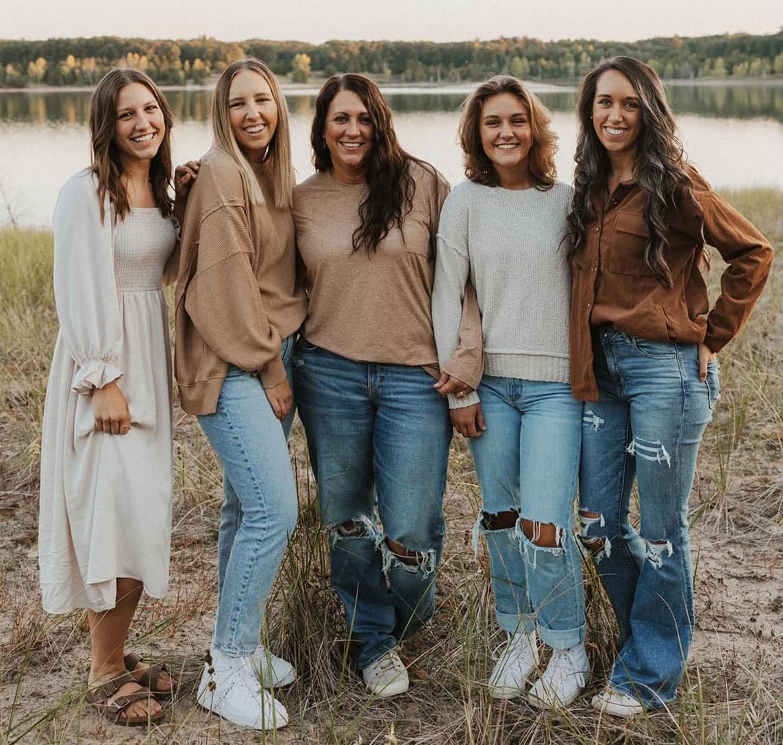 Group of five women outdoors near a lake, smiling and holding hands.