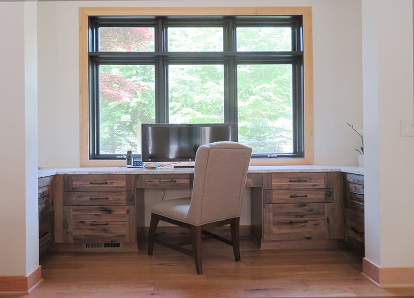 Home office desk with large window and natural light, wooden cabinetry, and a beige chair.
