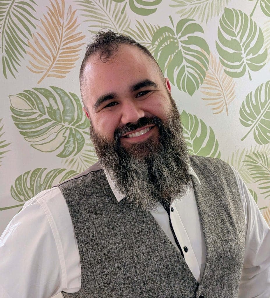 Smiling man with a beard in front of tropical leaf wallpaper.