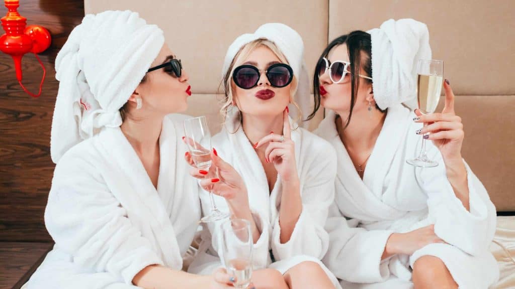 Women in robes and towels enjoying spa day with champagne and sunglasses.