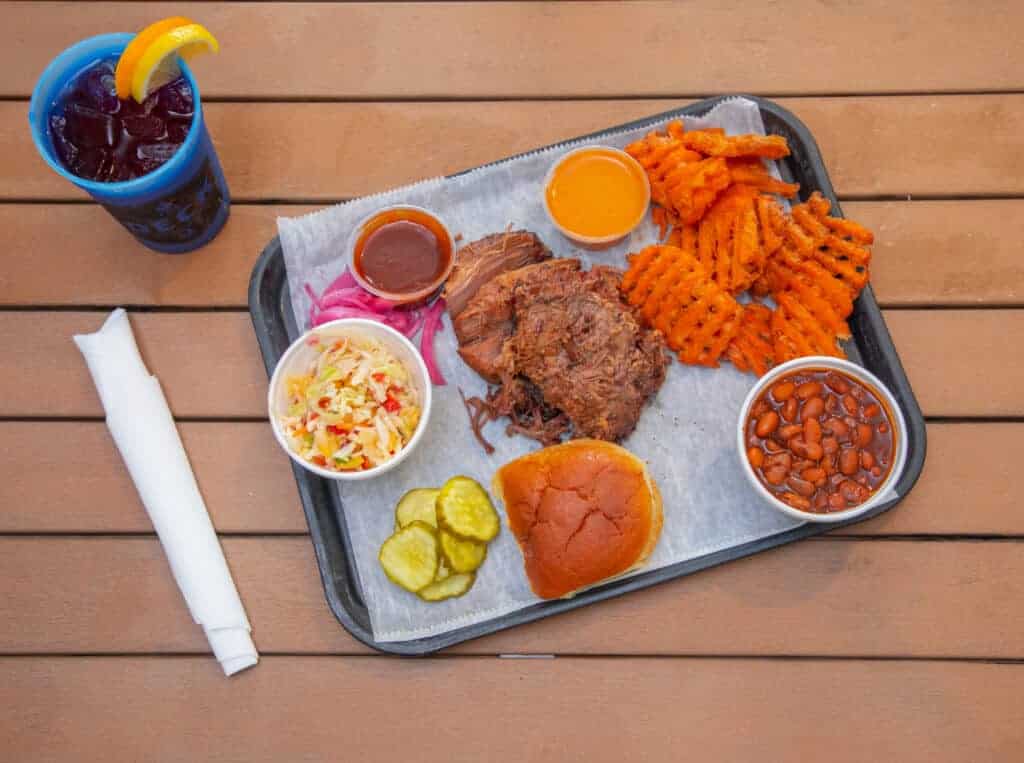 Meal with chopped brisket, waffle fries, baked beans, coleslaw, bun, and drink on wooden table.