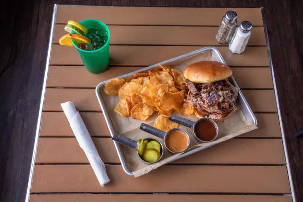 Juicy chopped brisket sandwich with chips, pickles, and drink on a metal tray.