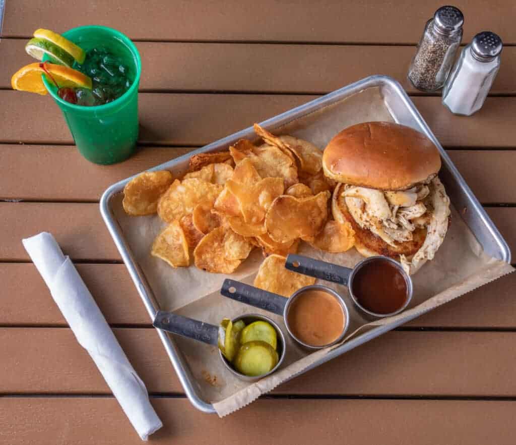Pulled smoked chicken sandwich with potato chips and dipping sauces on a tray.