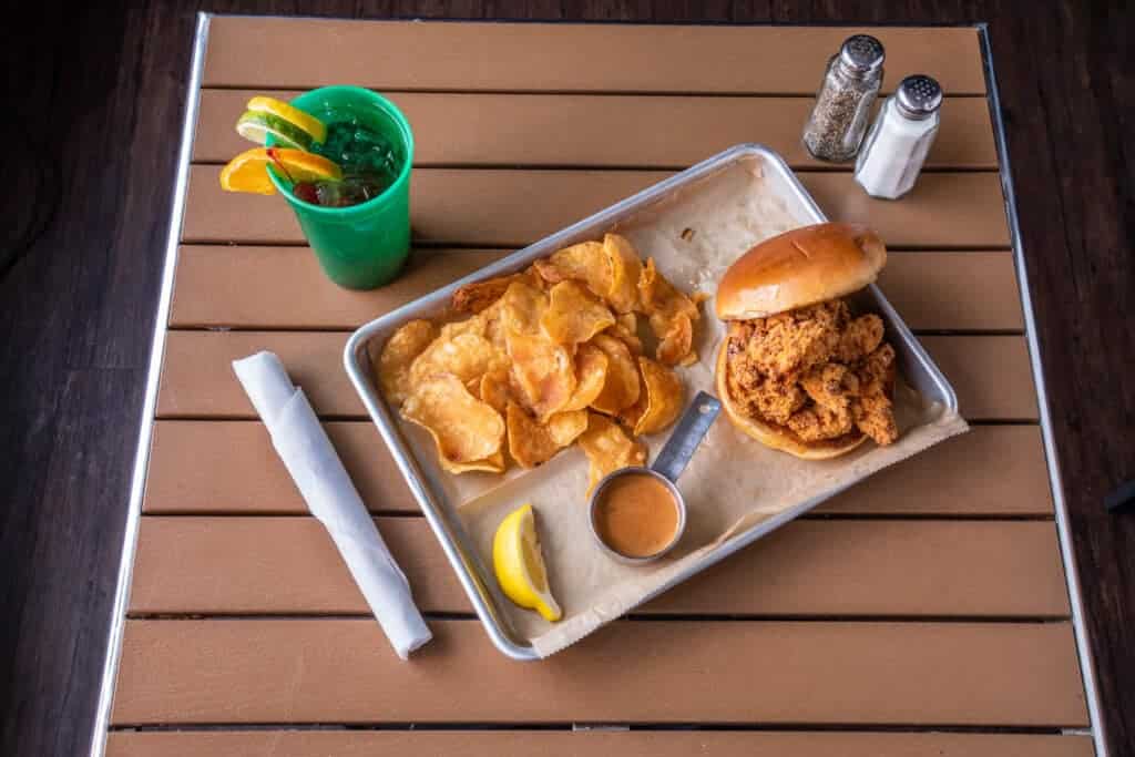 Crispy chicken sandwich with fried chips, lemon wedge, and drink on a tray.