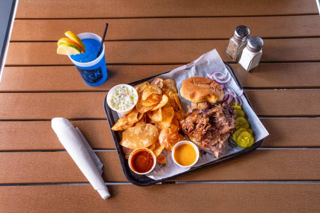 BBQ platter with fried chicken, potato chips, bread, and sauces on a wooden table.