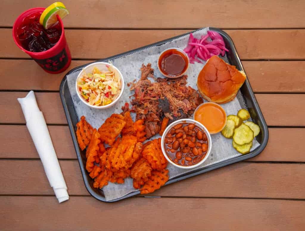 Pulled pork with baked beans, coleslaw, cornbread, and pickles on a tray.