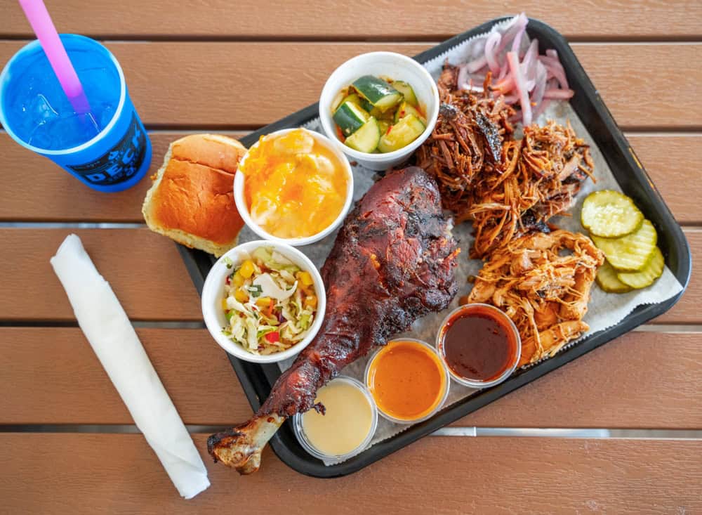 Barbecue platter with pulled pork, ribs, sides, and drink on wooden table.