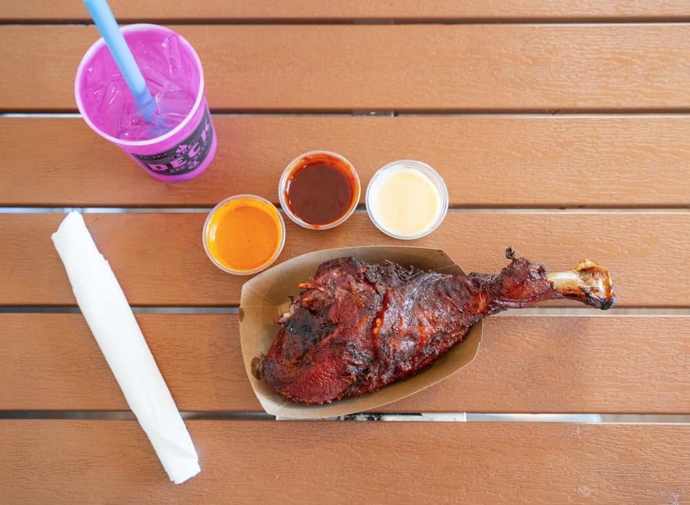 Juicy smoked turkey leg with three dipping sauces and a pink drink on wooden table.