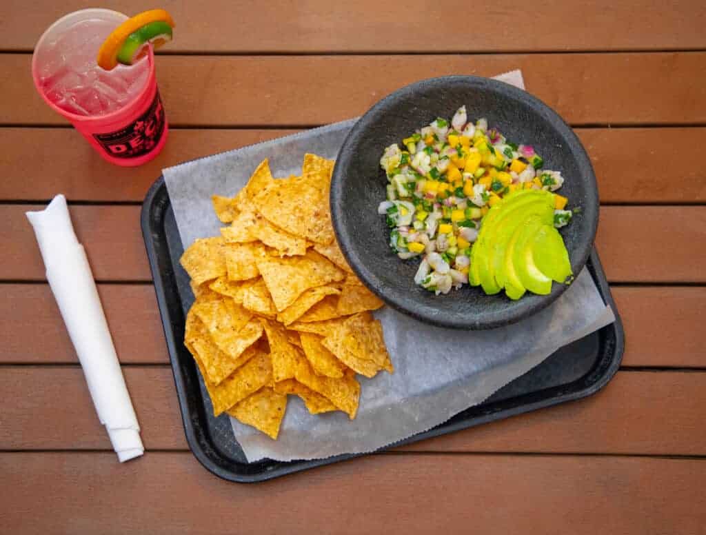Shrimp ceviche with avocado slices, tortilla chips, and a pink drink on a wooden table.