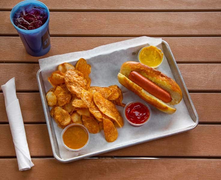 Hot dog with mustard, chips, and ketchup on a metal tray at The Deck.
