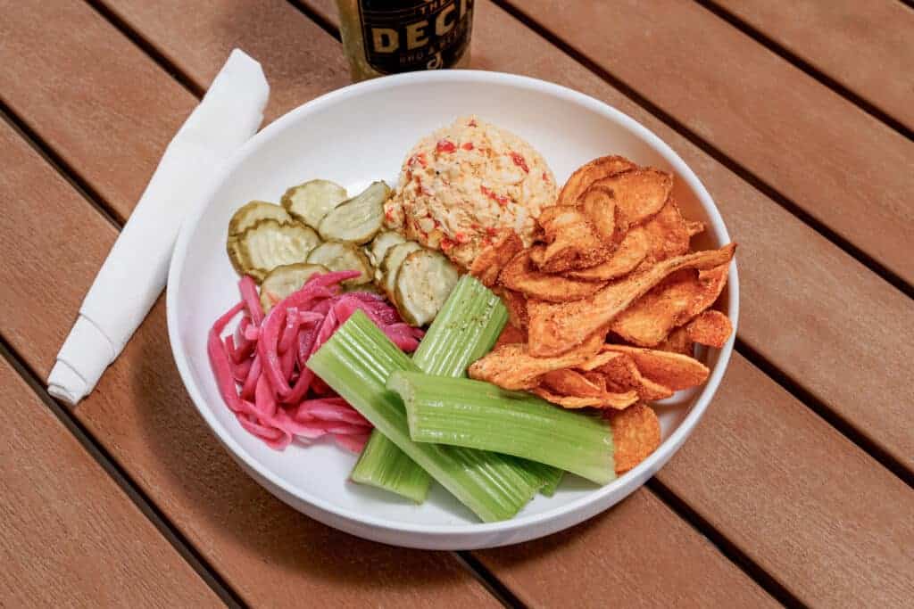 Pimento dip with pickles, celery, onion, and chips on a white plate.