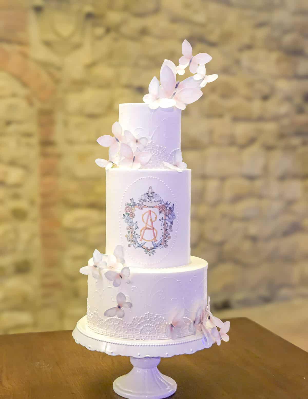 White tiered wedding cake with pastel monogram crest and pale pink sugar butterflies at Castello di Banfi in Tuscany, Italy.