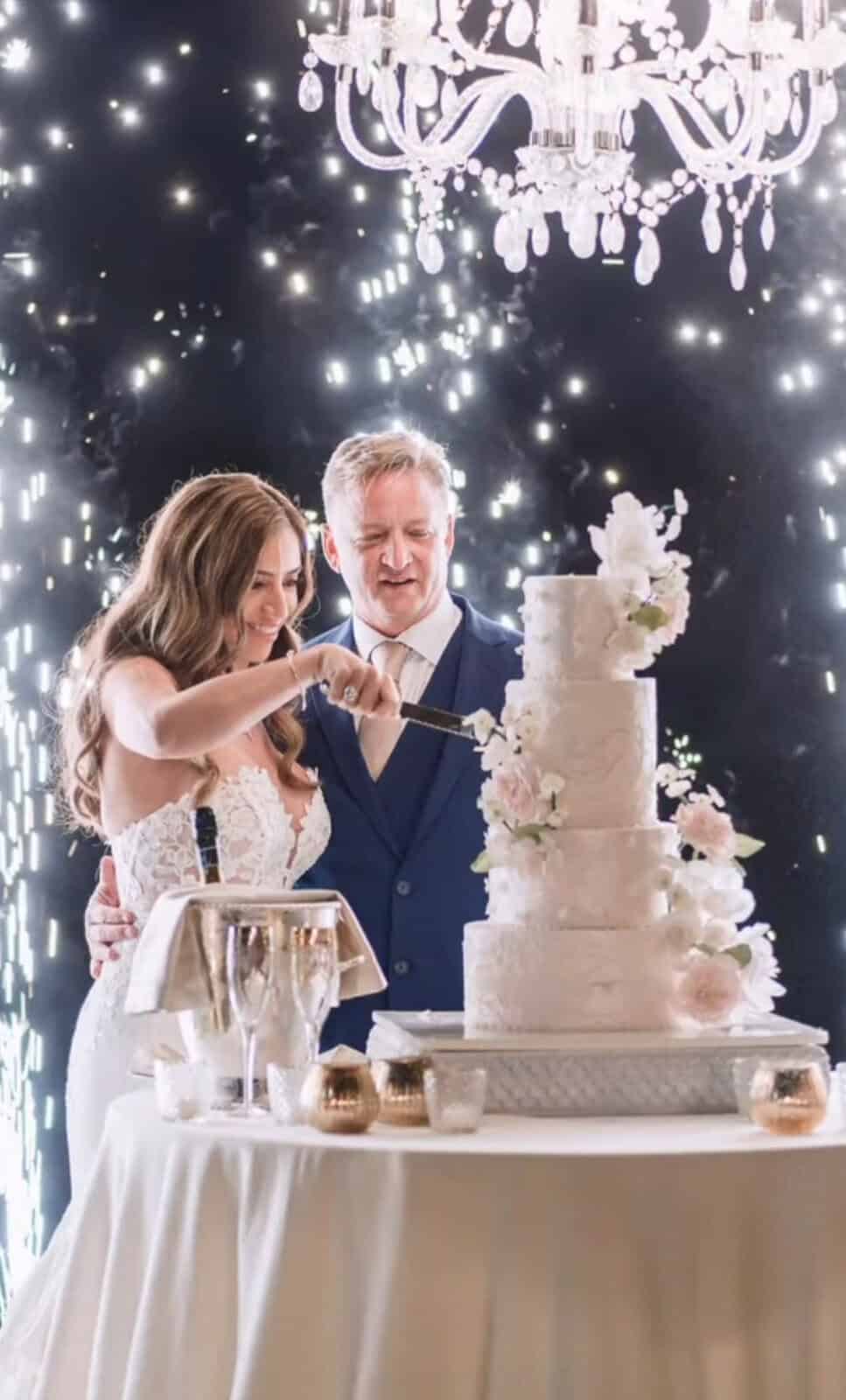 Couple cutting a tiered destination wedding cake in Italy under a chandelier with sparkler fireworks.