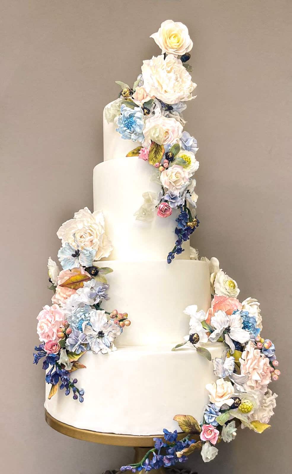 White tiered wedding cake with cascading sugar flowers in blue, blush, and ivory at Tuscany Bay Beach, Italy.