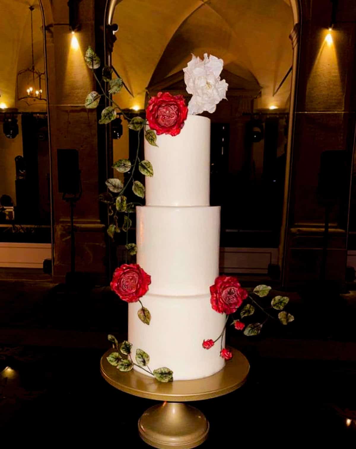 White tiered wedding cake with cascading red roses and greenery on gold stand at Villa San Michele in Fiesole, Tuscany, Italy.
