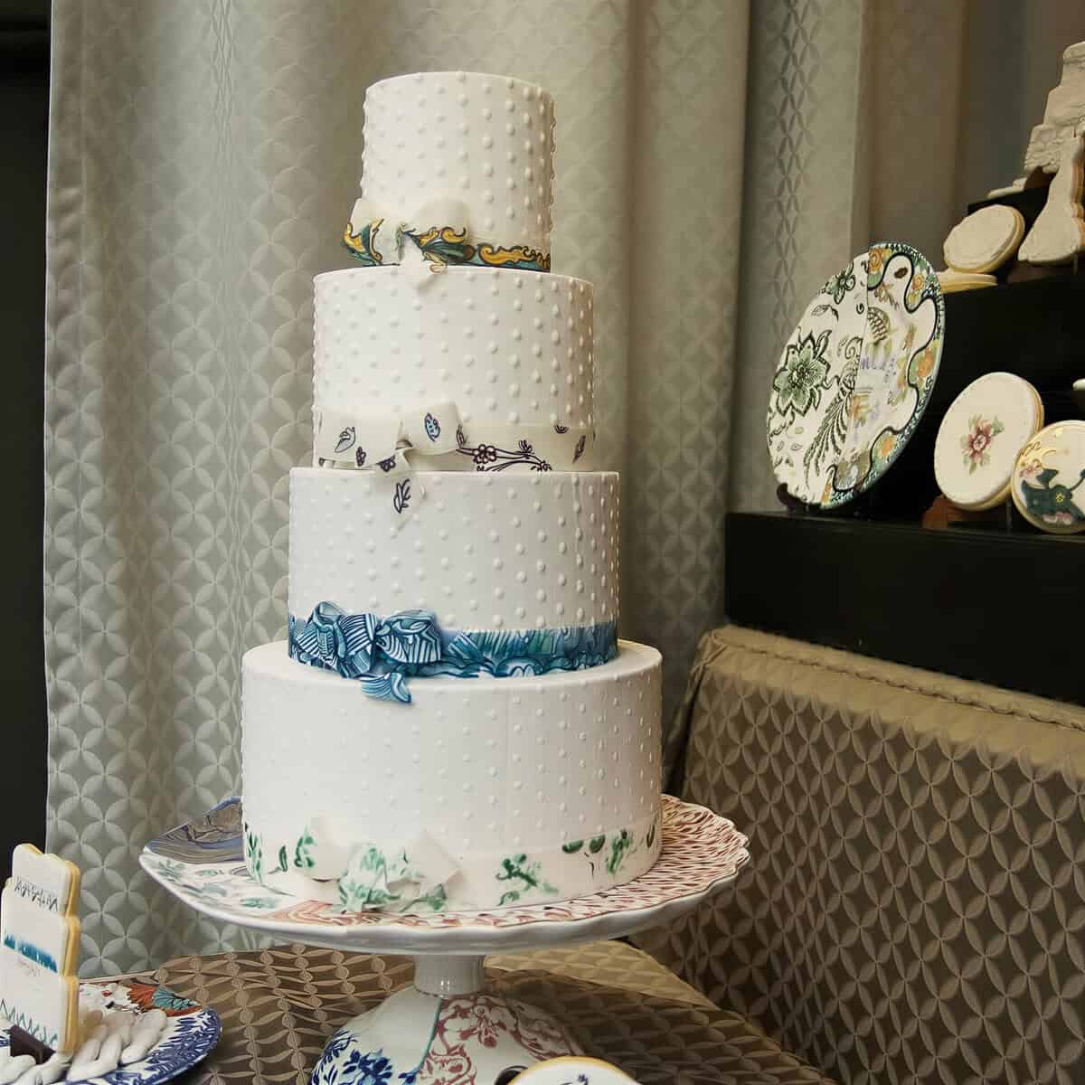 Four tier white cake with hand-piped dots and hand-painted sugar ribbons and bows in Seletti Hybrid plate patterns