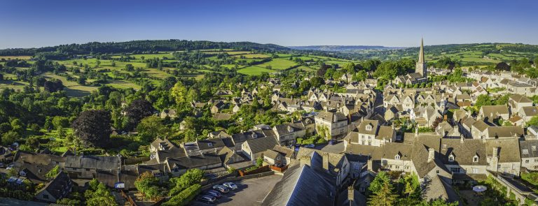 Clear blue summer skies and vibrant green patchwork fields above the iconic Cotswold village of Painswick, with its honey coloured limestone cottages and historic church spire. Representative of nature and biodiversity.