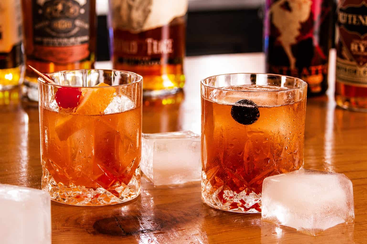 Two cocktail glasses with ice and drinks on a bar counter, with bottles in the background.
