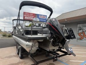 A pontoon boat with an outboard motor, displayed on a trailer outside a dealership, ideal for leisur.