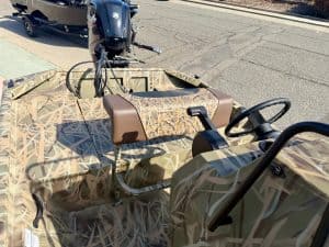 Camouflage boat with outboard motor, steering wheel, and seating on a city street.