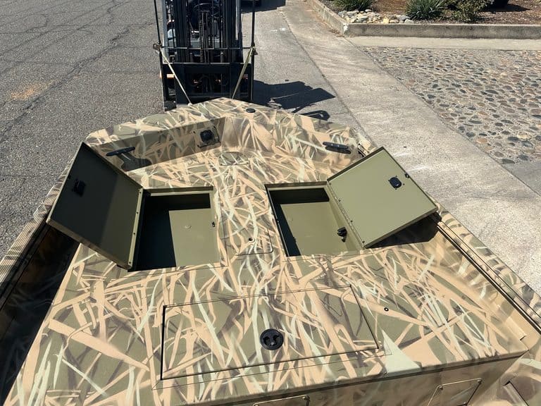 Camouflage boat with storage hatches and a forklift in the background.