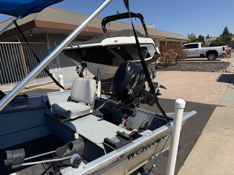 Small boat with outboard motor docked at marina with other boats and vehicles nearby.
