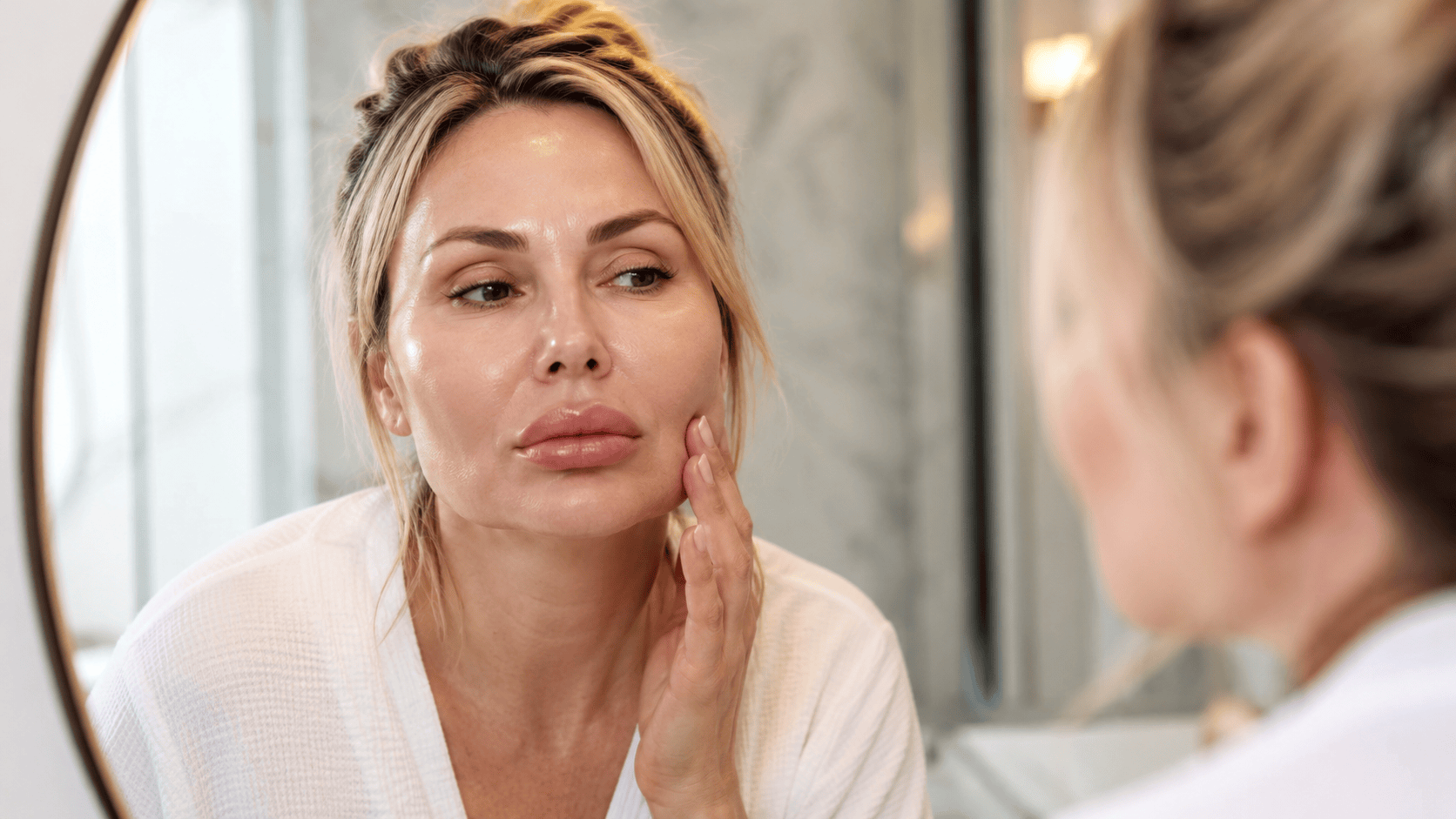 A woman studies her face in the mirror, checking whether her filler still looks natural.