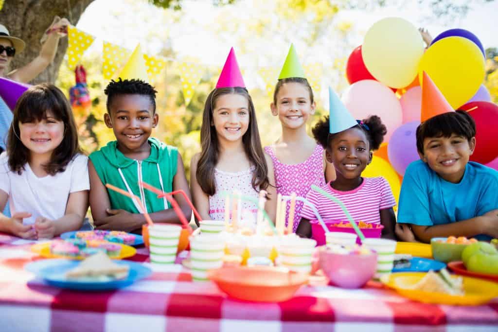 Kids celebrating birthday with cake, balloons, and party hats at an outdoor event.