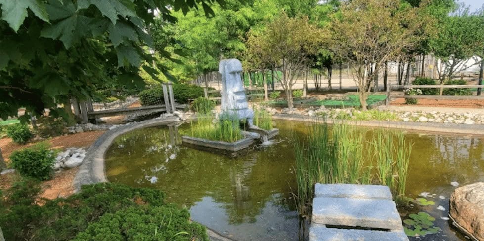 Garden pond with fountain, lush greenery, and trees in the background.
