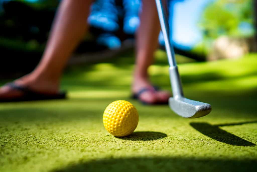 Close-up of a yellow golf ball on the green with a putter and a person in the background.