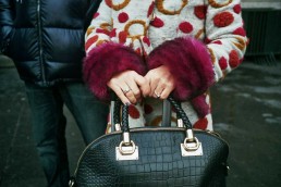 Closeup of the hands of a woman wearing a fancy coat with violet spots and fur carrying a black bag
