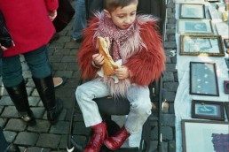 Street scene of a girl sitting in a baby carriage while eating a large sandwich in Brussels