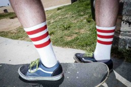 A foot of a man over a skateboard wearing white socks with 3 red horizontal stripes