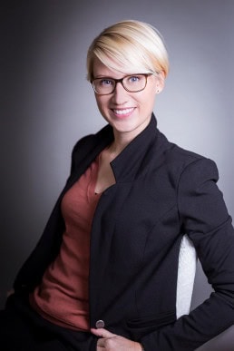 Corporate portrait of a young blond-hair woman with glasses in a suit against a dark background