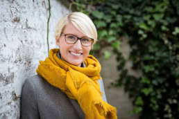 Portrait of a young blond-hair woman smiling wearing glasses, a grey jacket and a yellow scarf