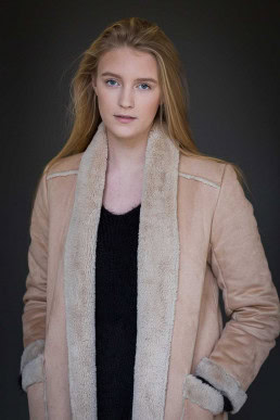 Headshot portrait of a blond woman wearing a beige coat against a dark background