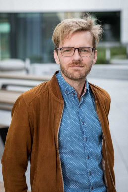 Portrait of a blond man wearing a blue shirt and brown jacket