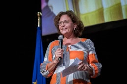 A woman smiling with a microphone introduces an award ceremony at Le Plaza Hotel in Brussels