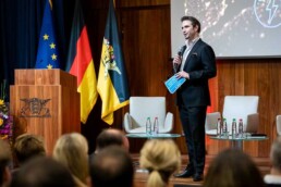 A host introduces an event and the European and German flags are on the background