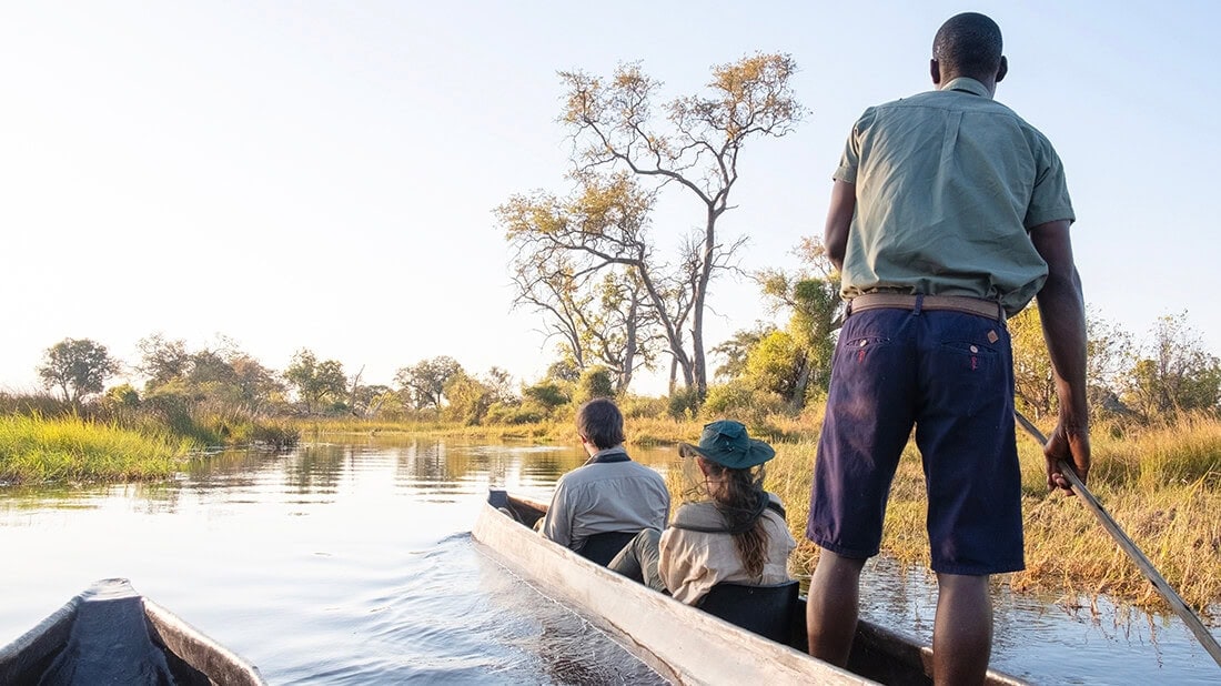 A couple sits with a guide in a mokoro as they travel down a river.