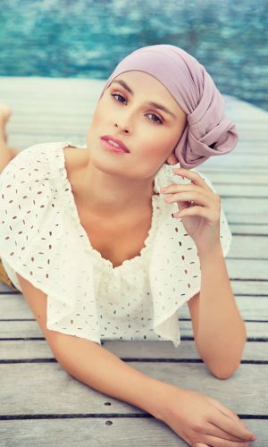 Woman with a light pink headscarf relaxing on a wooden dock by the water.