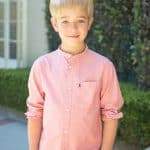 Young boy with blonde hair in a pink shirt outdoors, smiling at the camera.